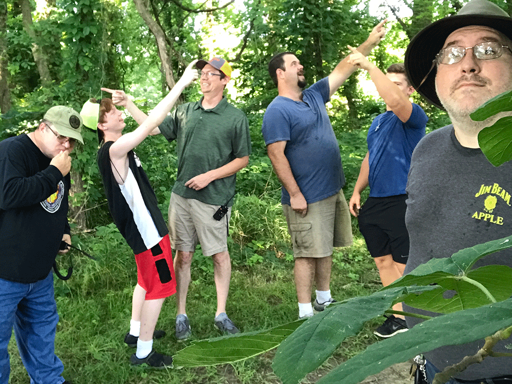 Six amateur radio operators stand among the trees and undergrowth, all of them pointing in different directions in a humorous manner.